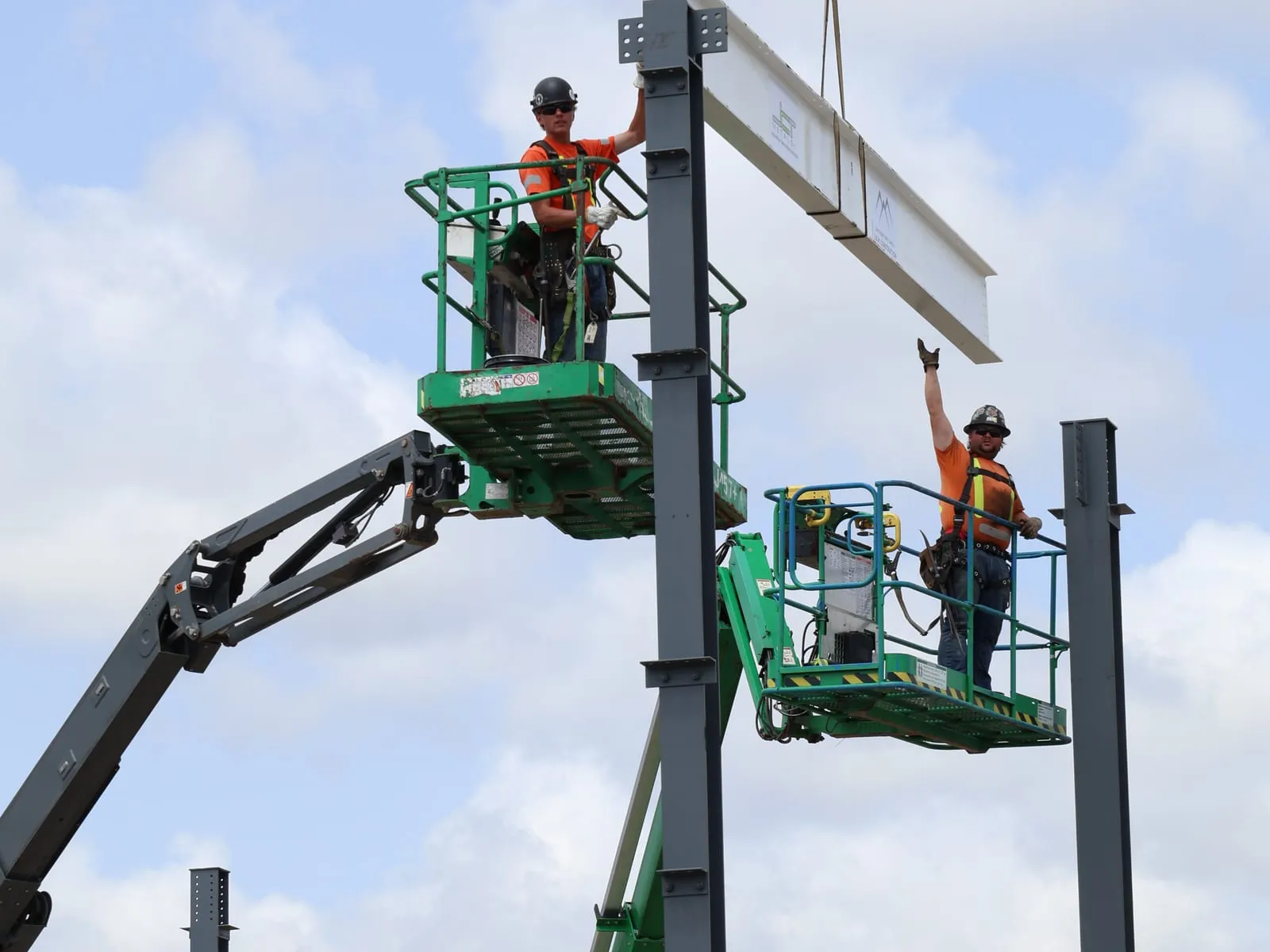 Two ironworkers in boom lifts guide a crane-hung steel beam onto the top of a column, against a cloudy sky.