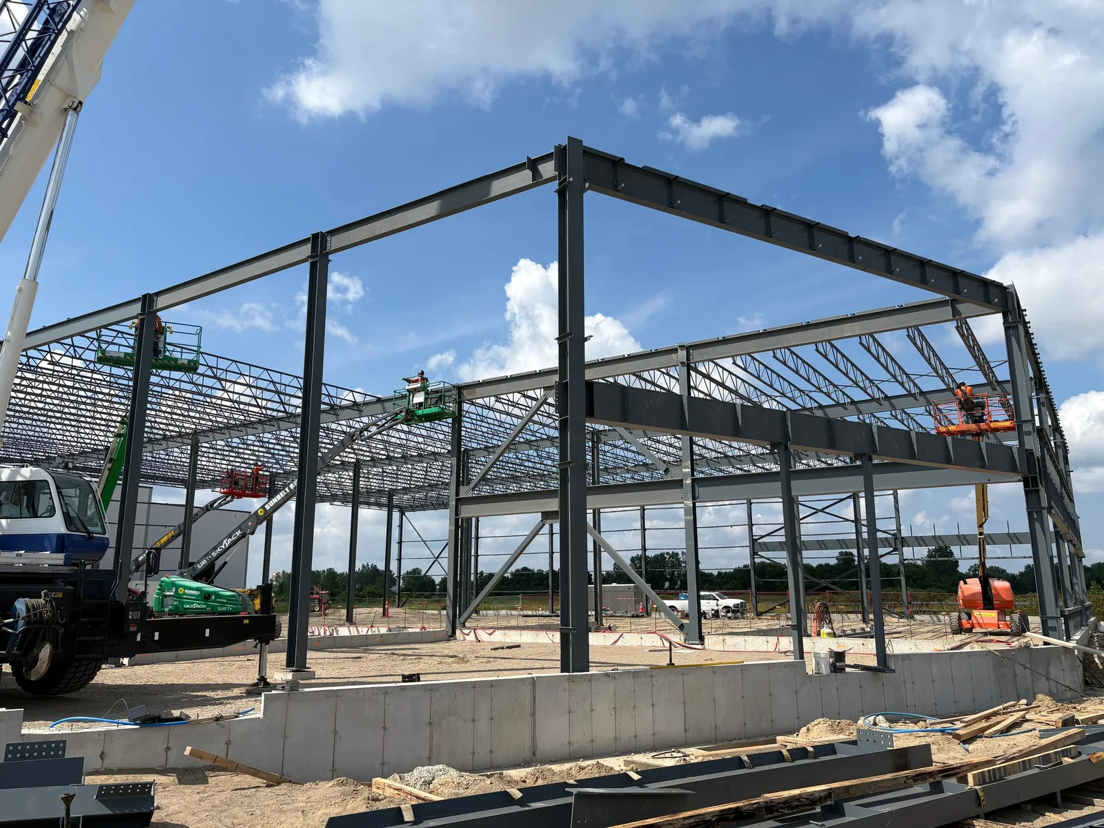 Ironworkers in boom lifts connect beams on a tall steel building frame under construction, with open-web roof joists behind, a mobile crane at the left and a concrete foundation wall in front.