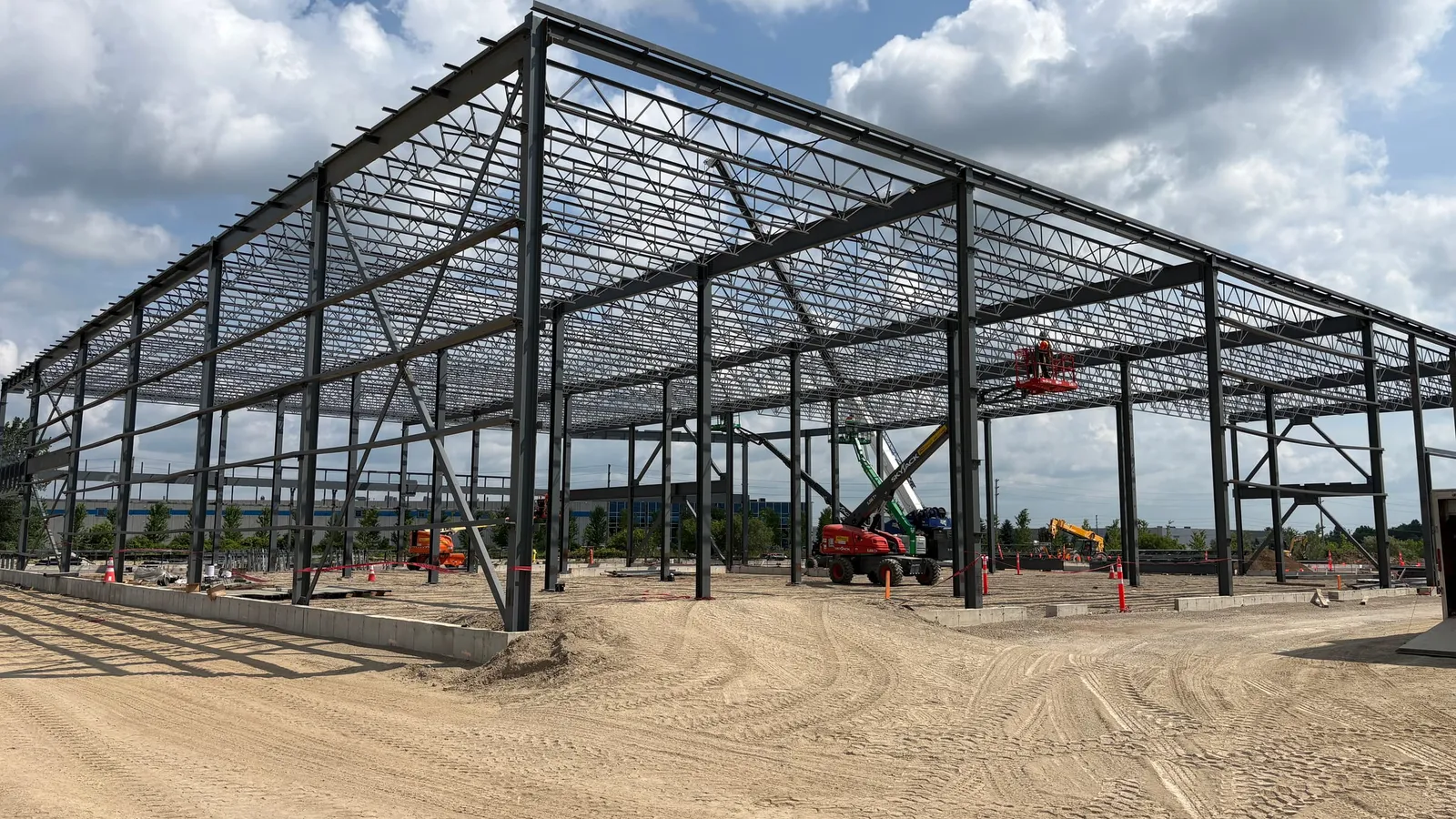 A large single-storey steel frame under construction: steel columns, beams and open-web roof joists, with no cladding yet, on a graded site.