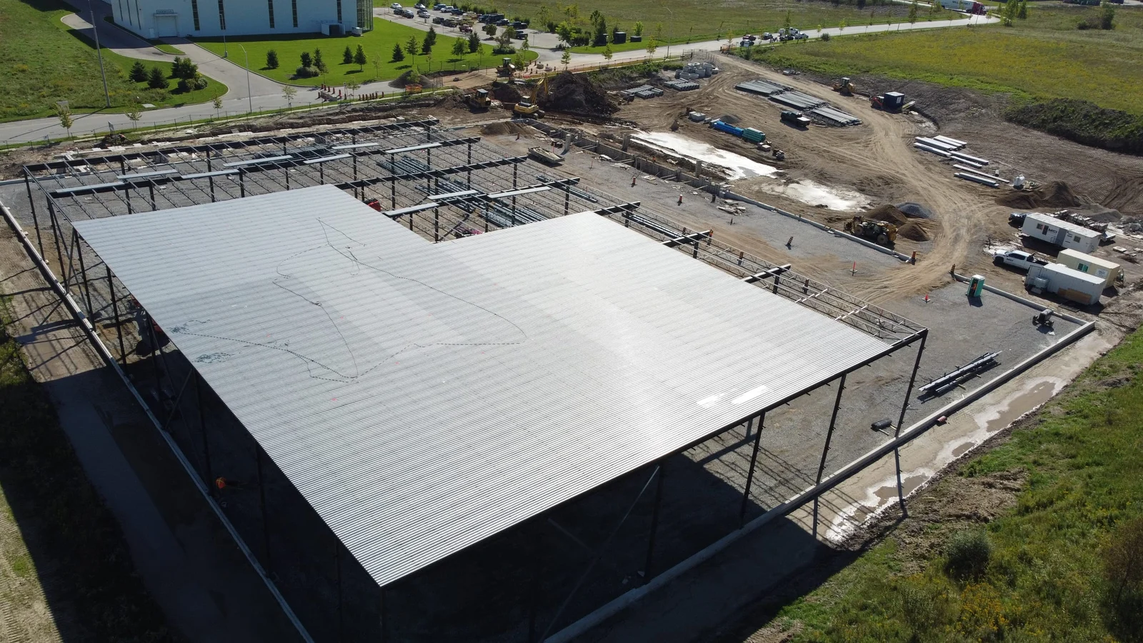 A high aerial view of a large steel building frame on a concrete slab, with an L-shaped area of silver roof deck already laid and green fields around the site.