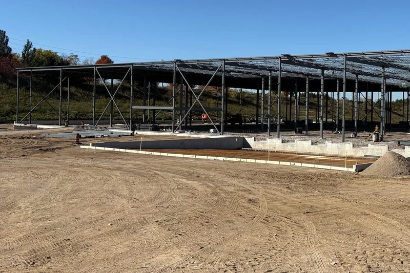 The long side of the braced steel frame across a graded site, with roof joists along the top and a concrete foundation wall in the foreground.