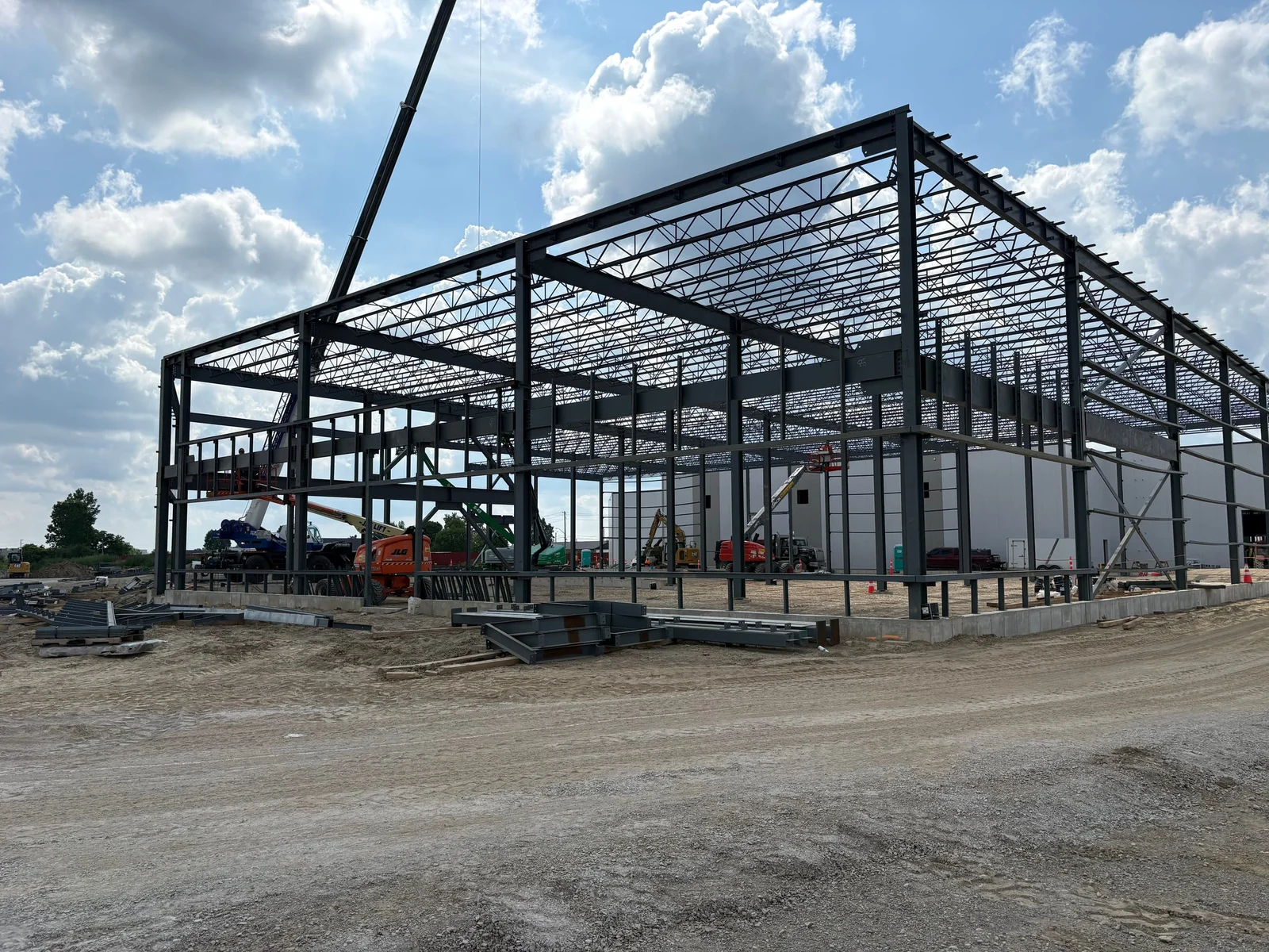 A tall single-storey steel frame under construction: columns, beams and open-web roof joists against a cloudy blue sky, with a crane boom at the left and steel members on the ground.
