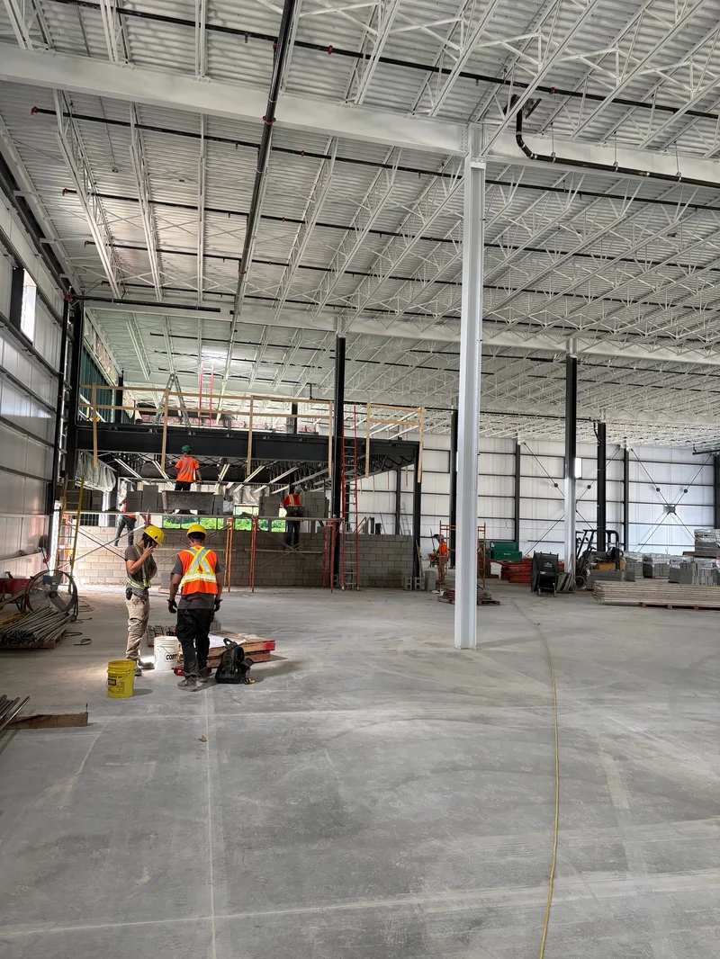 The Phase 1 interior during fit-out: white steel columns and an insulated roof on open-web joists, with masons on scaffolding at the mezzanine at the back.