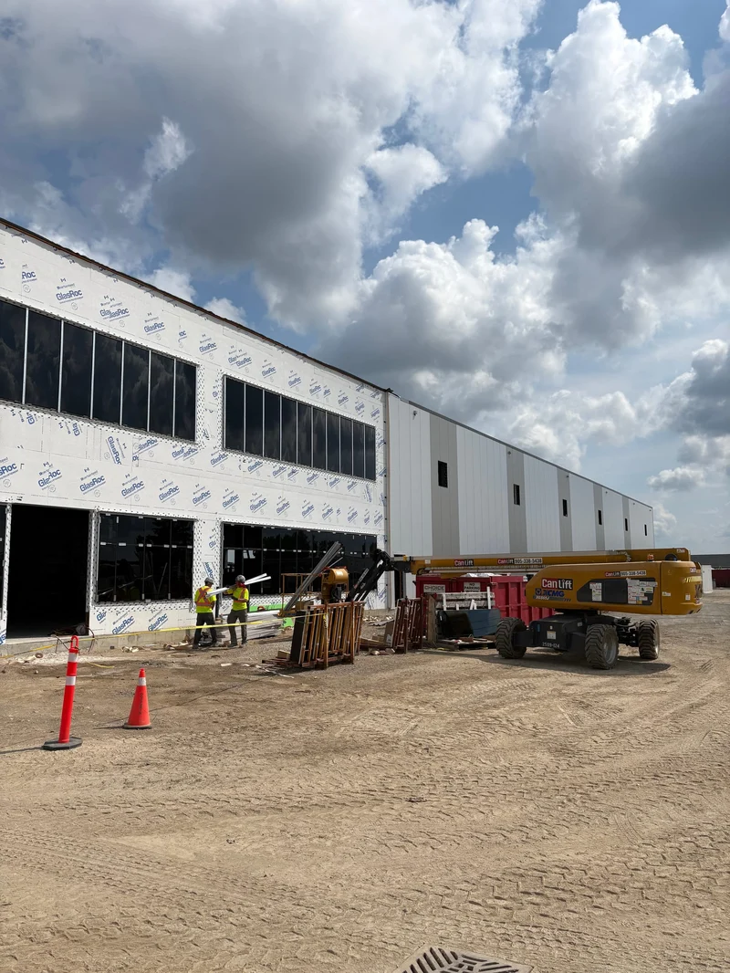 The clad front of the Phase 1 building, with rows of window openings in the two-storey office front, a boom lift and safety cones on the gravel in front.
