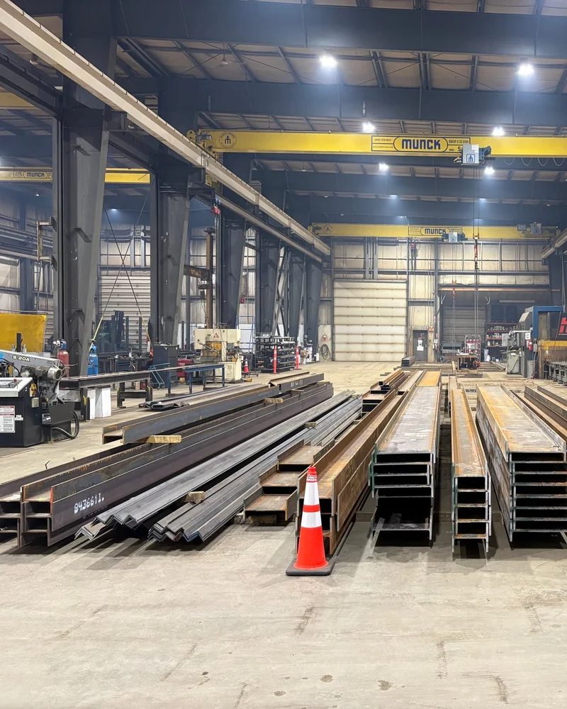 A long shop bay with wide-flange beams and steel angles laid out on the floor beside an orange safety cone, under yellow overhead cranes.