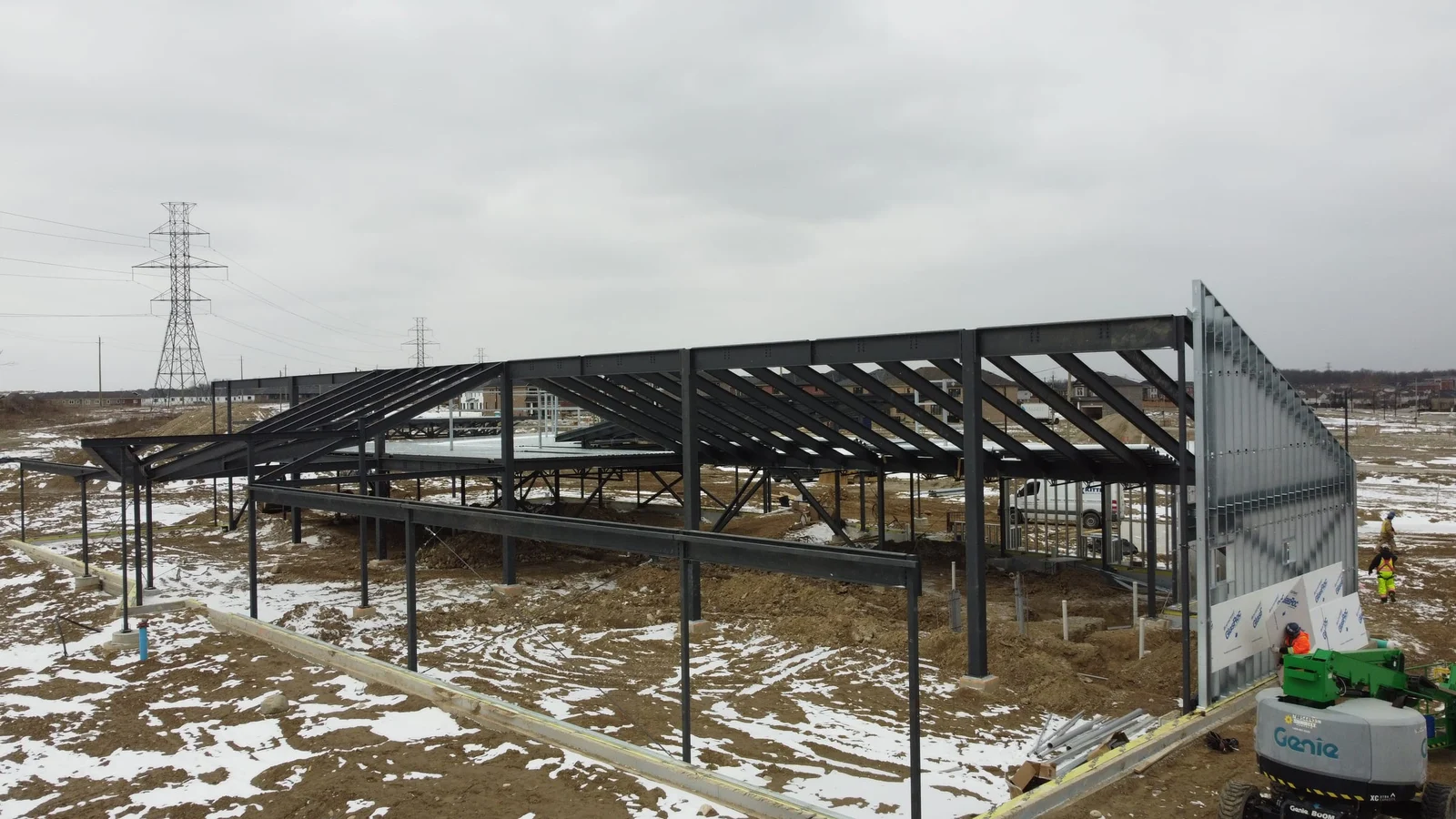 An aerial view of a library under construction in winter: sloped steel roof framing cantilevering past the walls, a tall steel-stud gable wall and a worker in a boom lift.