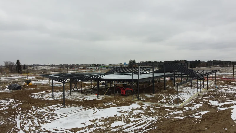 A wide aerial view of the whole steel frame on a snowy site, with vehicles and building materials around it.