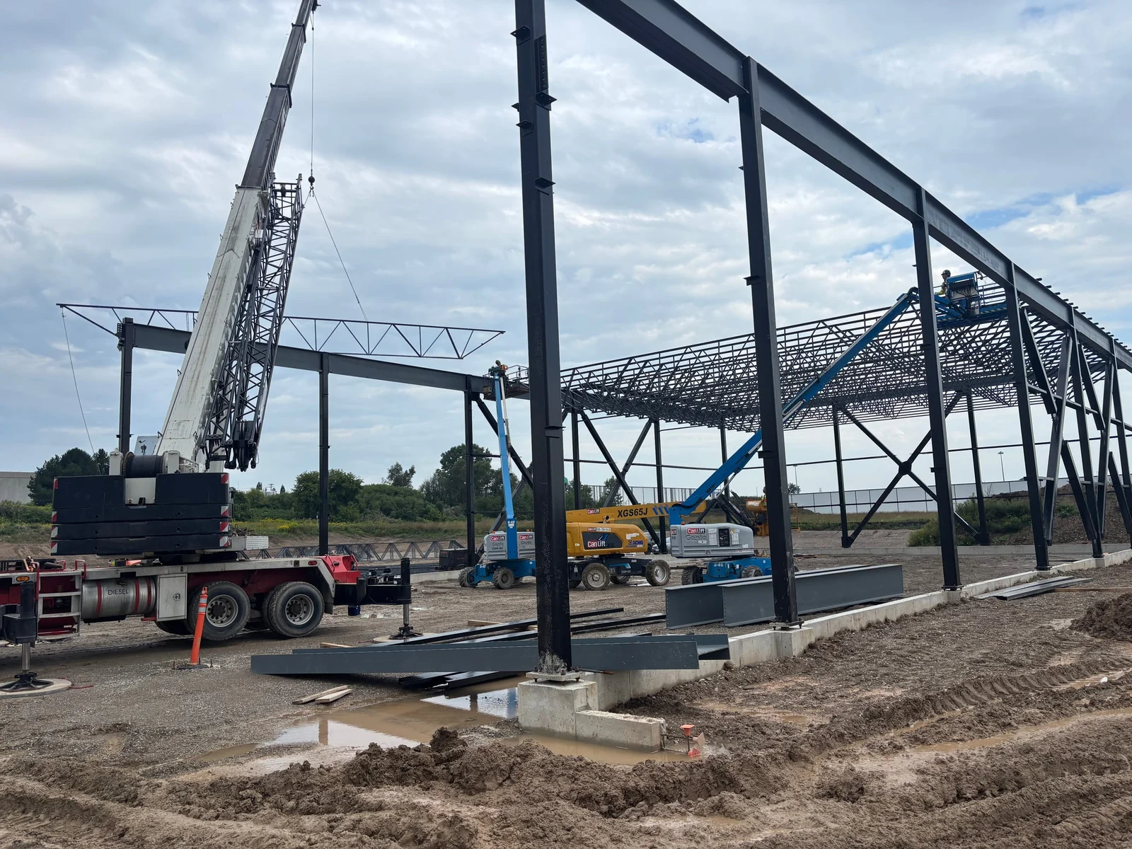 A mobile crane and boom lifts beside tall steel columns and a braced frame, with open-web roof joists set at the back of the site.