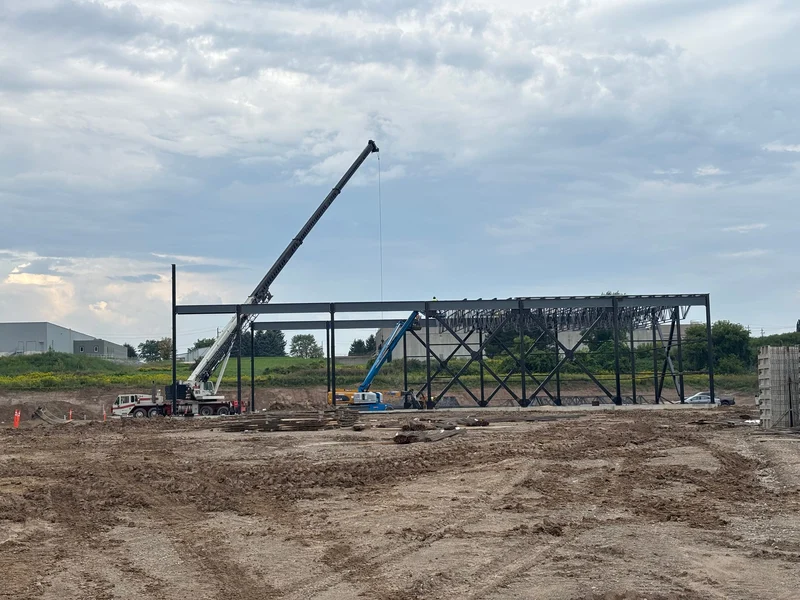 A long braced steel frame across a muddy site under an overcast sky, with a crane lifting steel at the left and a boom lift beside the frame.