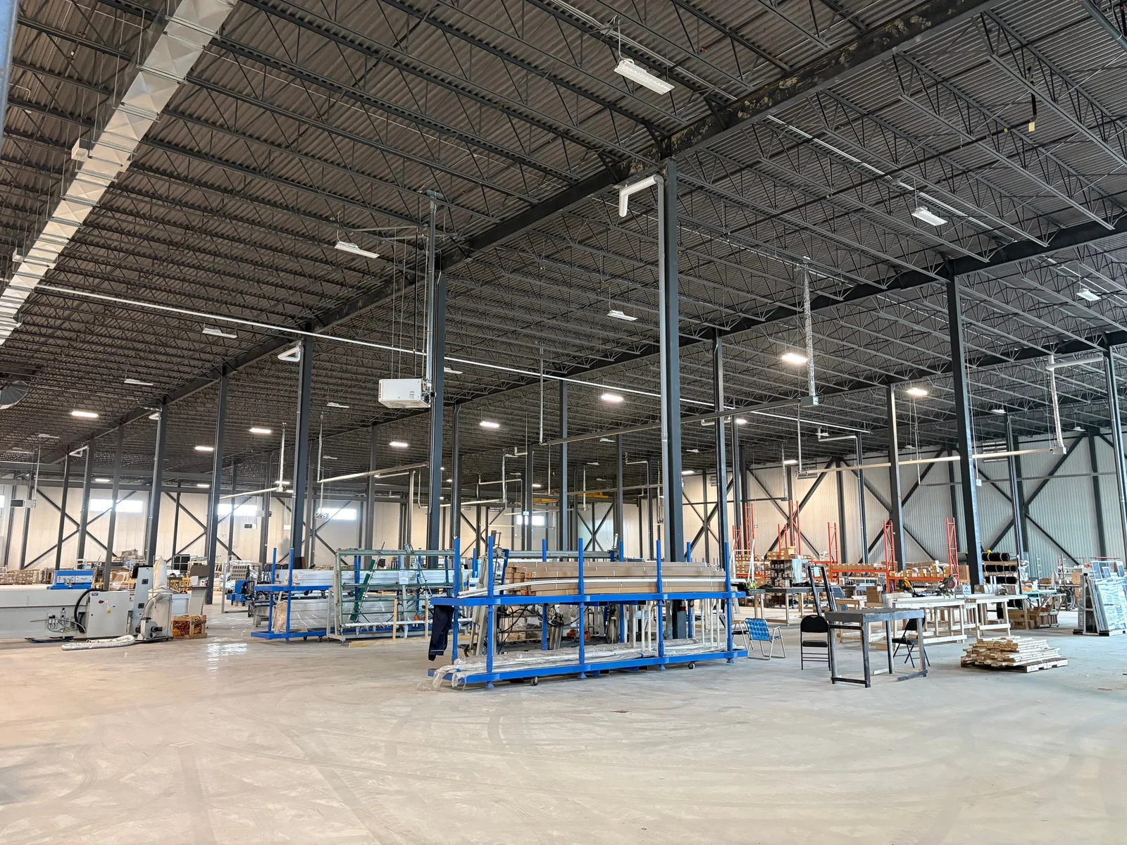 The finished interior of a large industrial building: a high roof on open-web joists over steel columns and X-bracing, with a production line being set up on the floor.