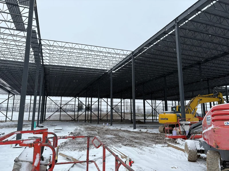 A huge field of open-web roof joists on steel columns going up over snow-covered ground, with an excavator and a boom lift on site.