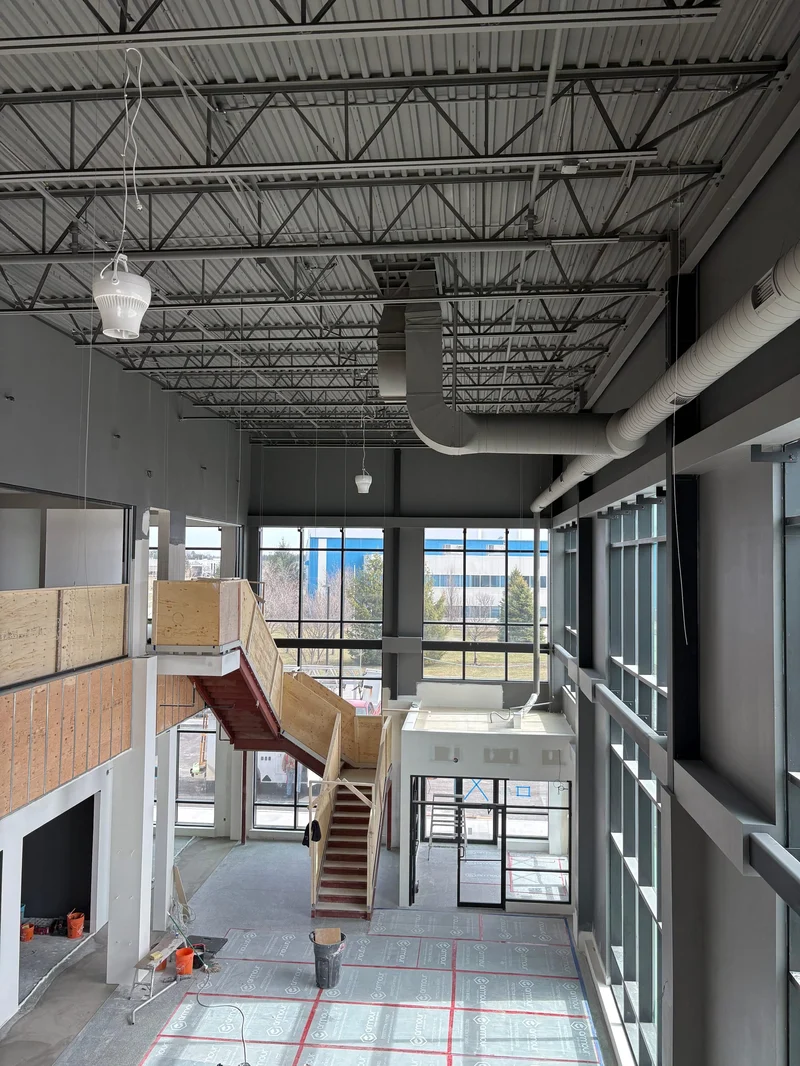 A two-storey office atrium under construction: a curtain wall, a steel stair boxed in plywood guards, floor protection, and exposed steel joists above.