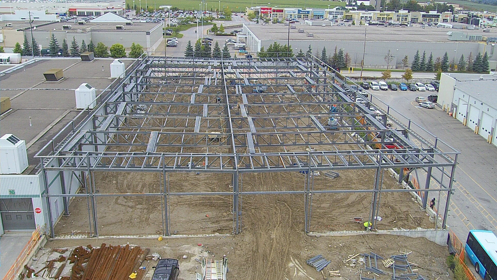 An aerial view of a large multi-bay steel frame beside an existing bus garage, with steel columns, beams and roof joists in place and rebar bundles on the ground.