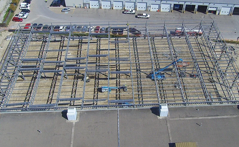 Looking down on the steel frame from above: rows of open-web joists between beams, with boom lifts working and parked buses along the garage.